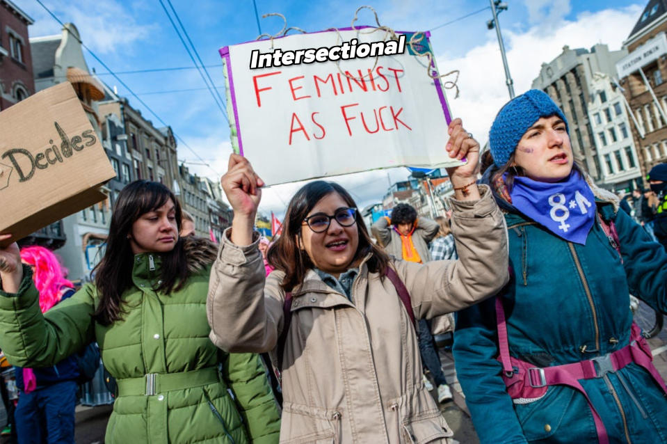 Three individuals at a rally holding a sign that reads "Feminist as F***" expressing advocacy for women's rights