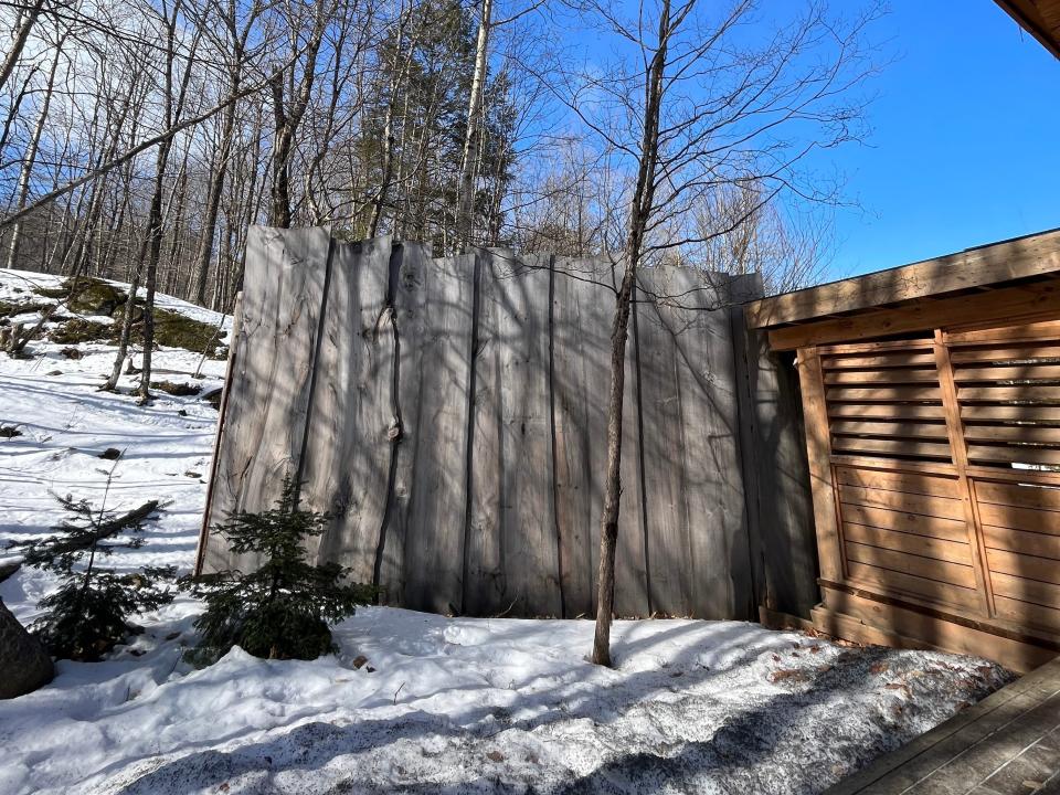 The side of a wooden lodge with a tall wooden fence surrounded by trees and snowy ground