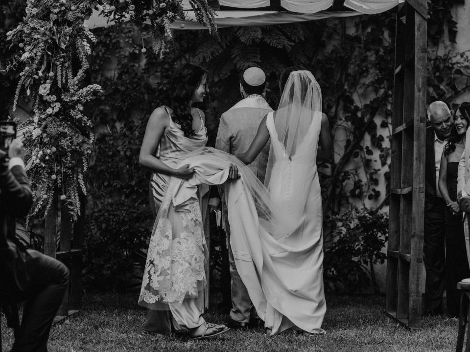 A bride and groom stand under an archyway as a woman holds the train of the bride's dress.