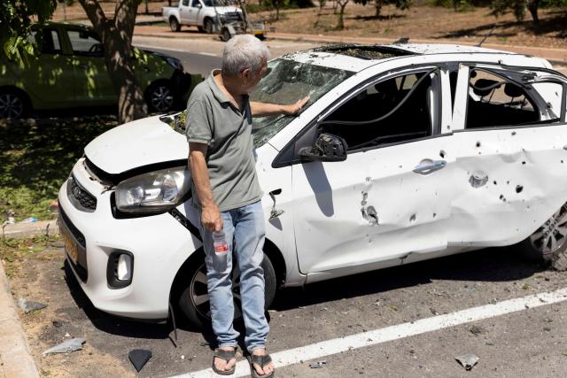 A man stands next to his wrecked car which has been hit by shrapnel 