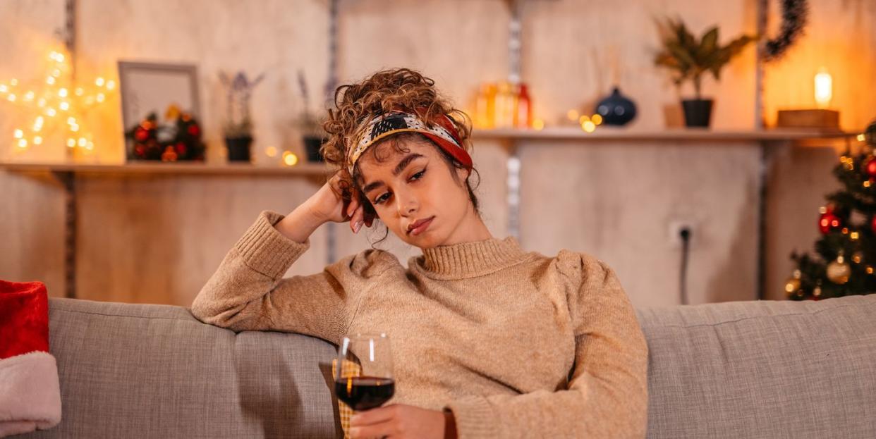 lonely and sad young woman drinking a glass of red wine alone on the sofa with christmas lights and a tree behind her