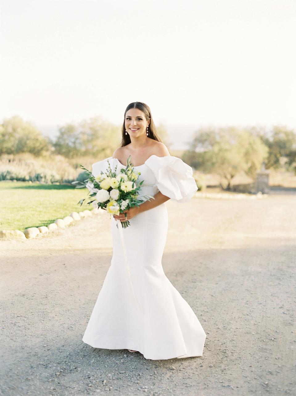 A bride stands in her wedding dress holding a bouquet of white and yellow flowers.