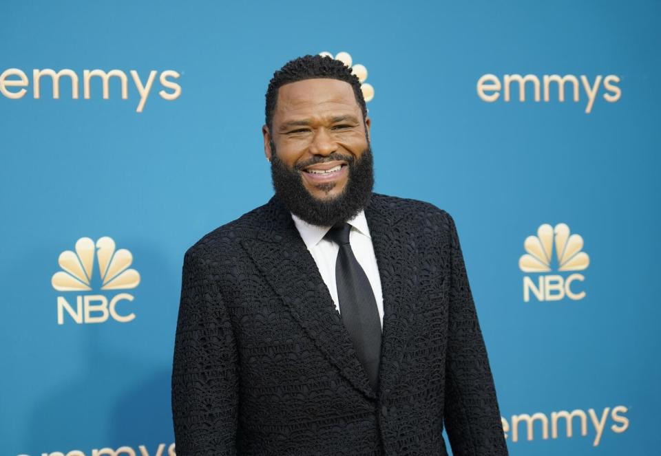 Anthony Anderson in a black suit and tie stands in front of a blue backdrop