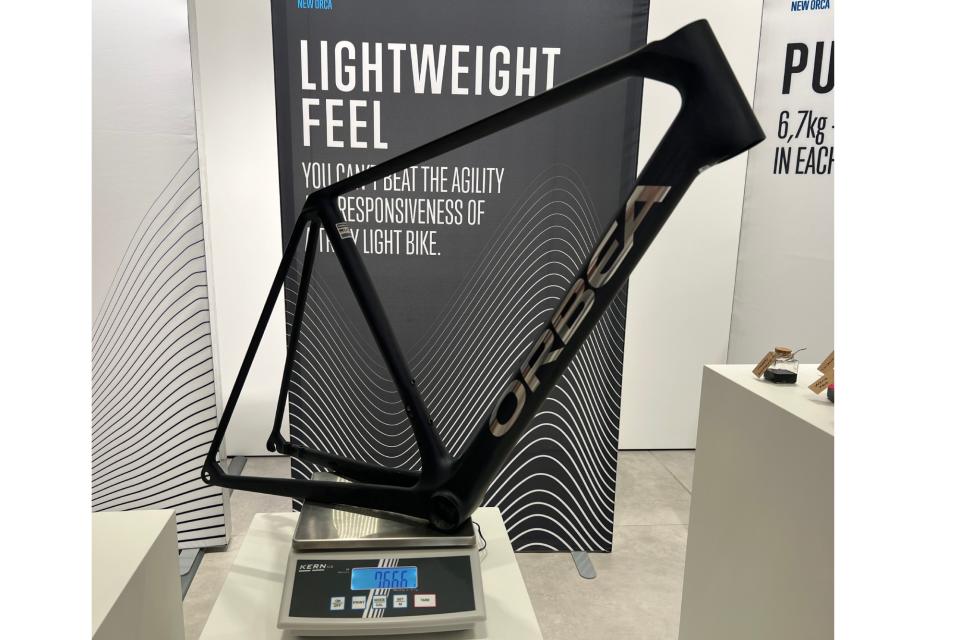 The Orbea Orca OMX frame balanced on some table top scales with a poster in the background which reads Lightweight feel as a headline