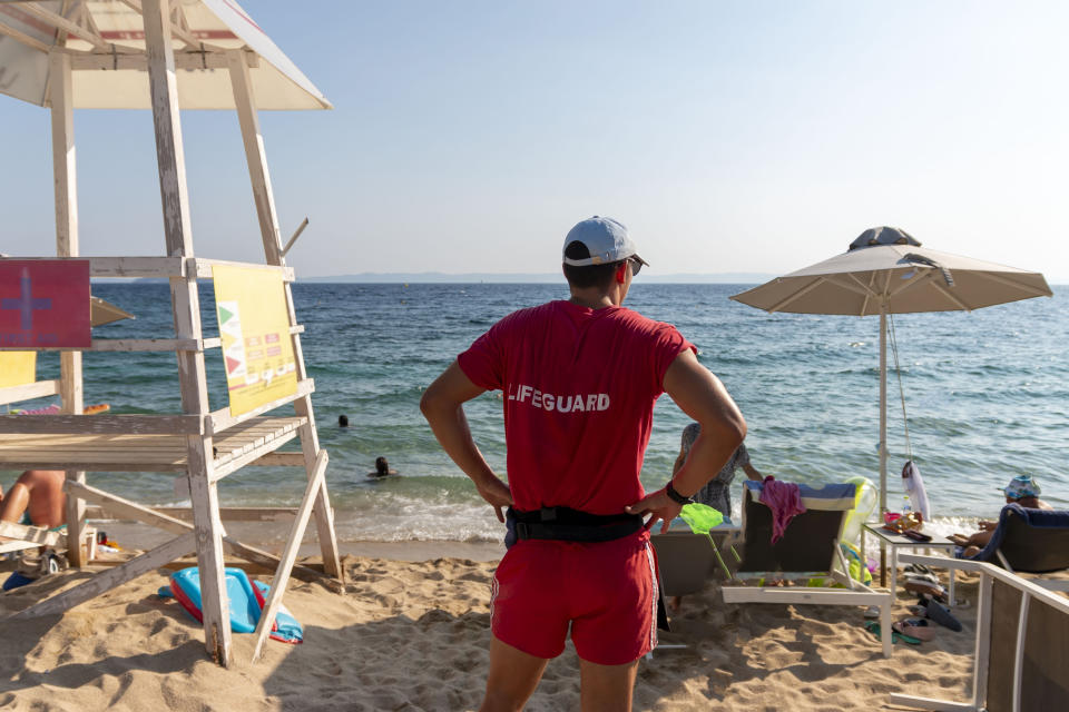 A lifeguard stands on a sandy beach, overlooking swimmers in the sea. Deck chairs and an umbrella are visible near the lifeguard tower