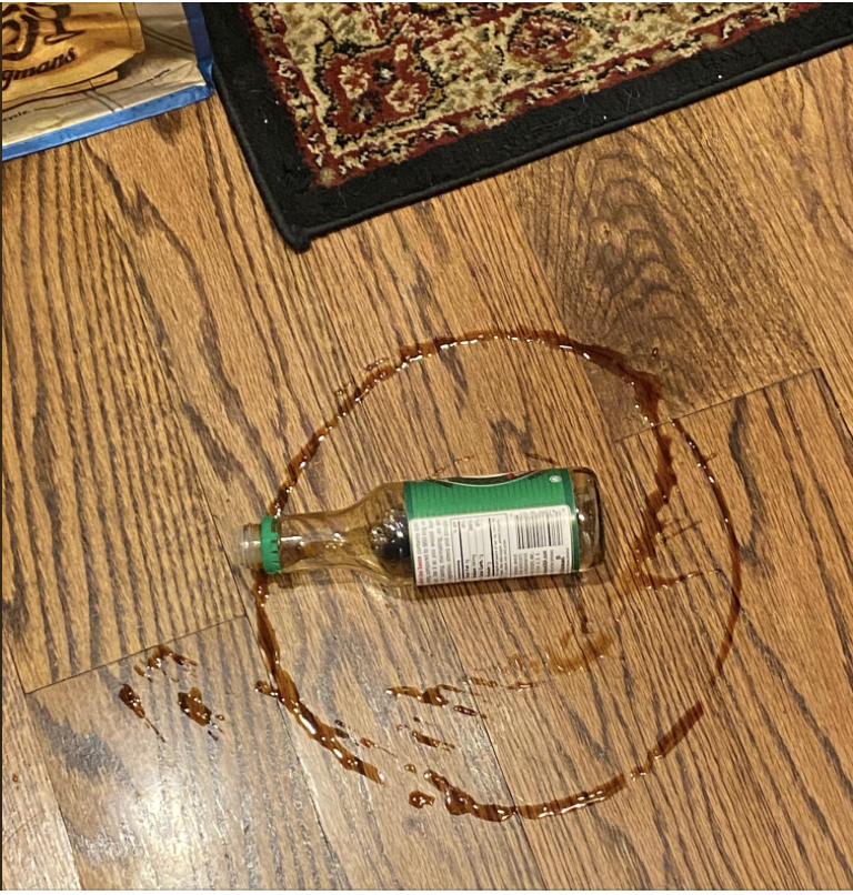 A bottle of soy sauce lies on a wooden floor with a spill around it