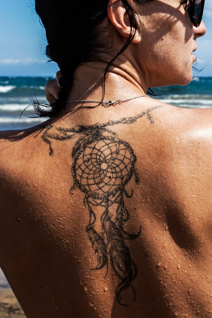 Person with a dreamcatcher back tattoo looking at a beach with kitesurfers in the distance