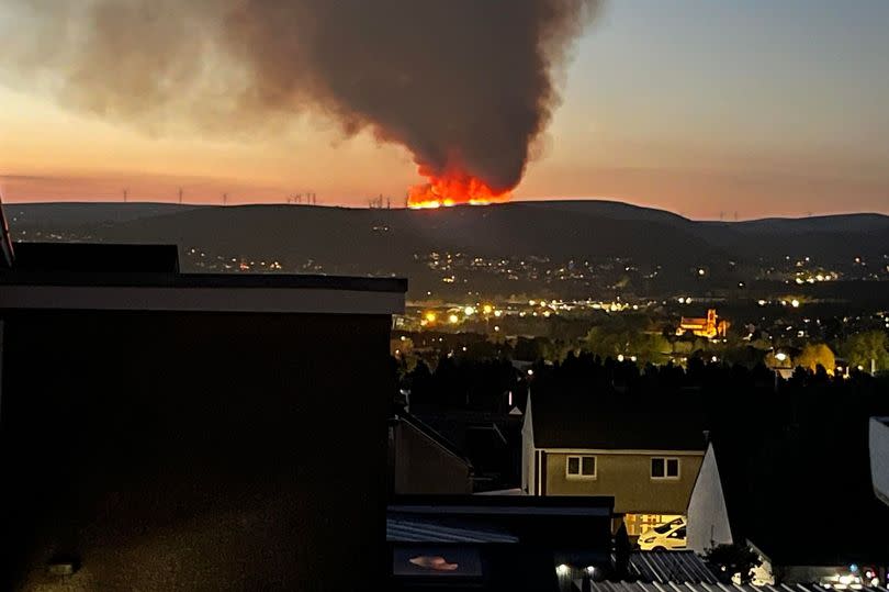 A fire rages in the distance on mountain, with houses visible in foreground