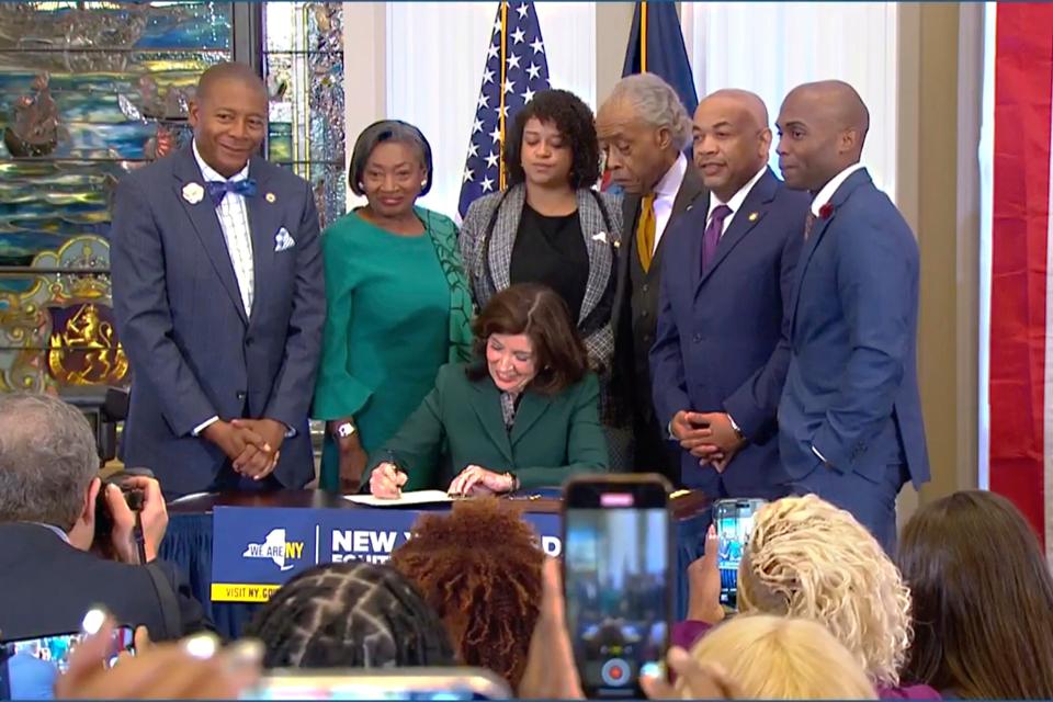 This image from video provided by the Office of The Governor, shows New York Gov. Kathy Hochul signing a bill in Albany, NY, Tuesday, Dec. 19, 2023, to create a commission tasked with considering reparations to address the persistent, harmful effects of slavery in the state. She is joined by, standing from left: Dr. Yohuru Williams, Founding Director of the Racial Justice Initiative at the University of St. Thomas; Andrea Stewart-Cousins, Majority Leader of the NY State Senate; Michaelle Solages, NY State Assemblymember; Rev. Al Sharpton; Carl Heastie, Speaker of the NY State Assembly; James Sanders, NY State Senator. (Office of the Governor via AP)