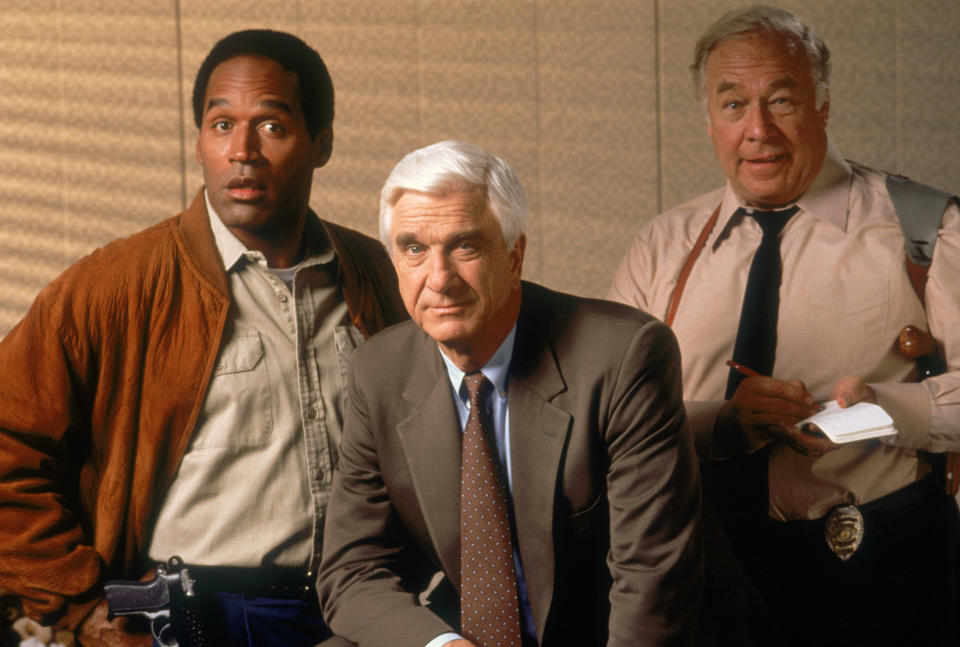 Three actors on a film set, one wearing a leather jacket, a second in a suit, and a third in a shirt, with props suggesting they are playing police officers