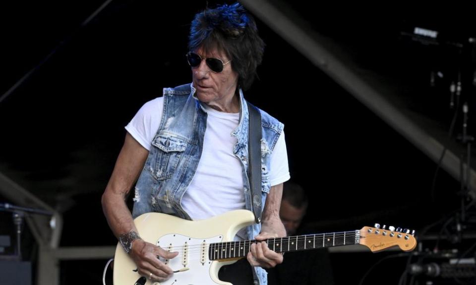 Beck plays guitar on stage, in white t-shirt and sleeveless denim jacket.