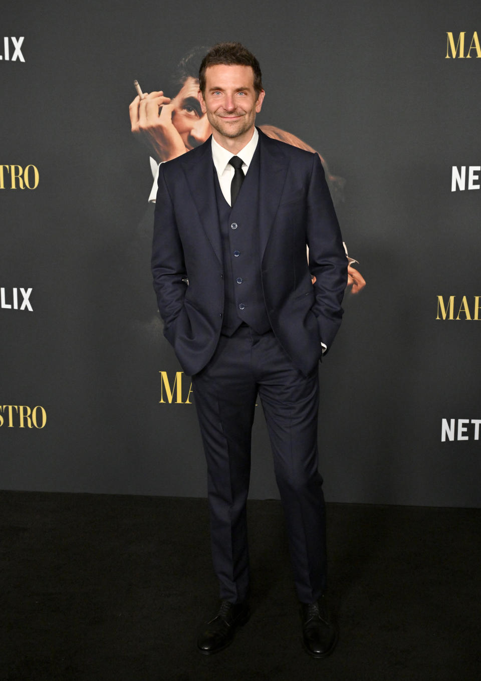 Bradley Cooper stands smiling in a tailored navy suit with a tie, at a Netflix event for "Maestro"