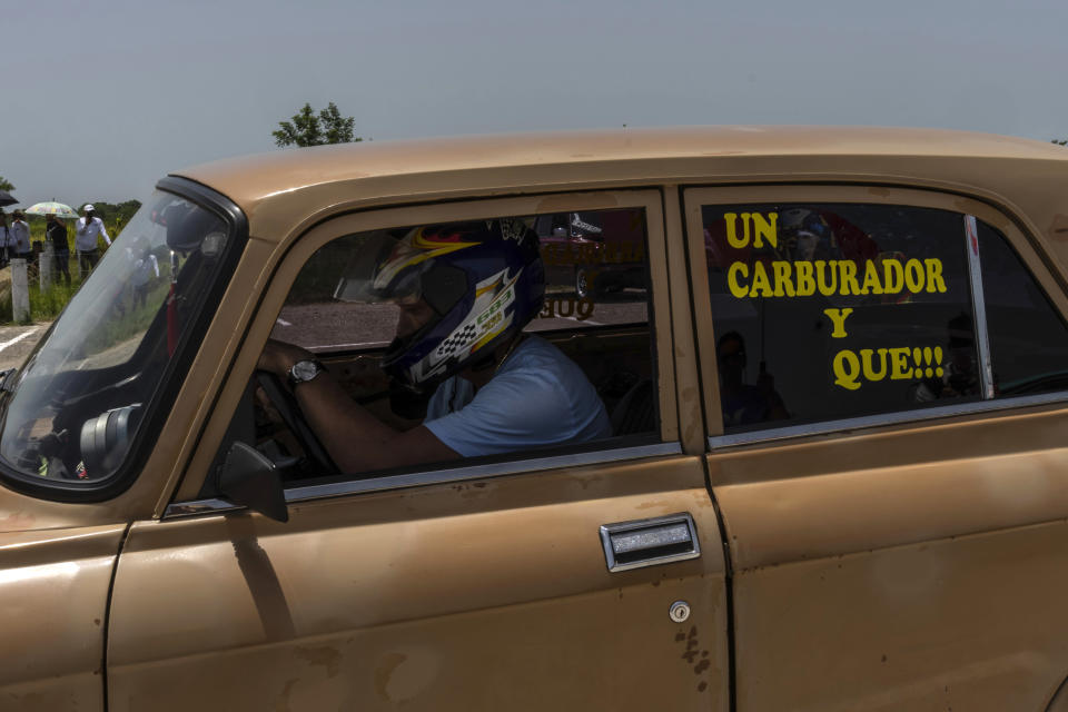 Un conductor usa un casco mientras prueba el arranque de su viejo auto Moskvitch antes de una carrera de resistencia en una vieja pista de aterrizaje sin usar en San Nicolás de Bari, Cuba, el domingo 23 de julio de 2023. El mensaje en la ventana dice en español "¡Un carburador y qué!", refiriéndose a la broma de un mecánico que cuestiona de qué otra cosa está hecho el auto. (Foto AP/Ramón Espinosa)