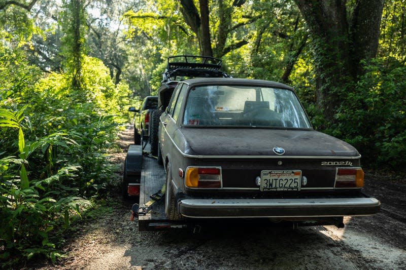 a 1974 bmw 2002tii shown on a car trailer being towed through a narrow passage in an overgrown field, surrounded by lush trees and bushes somewhere in florida.