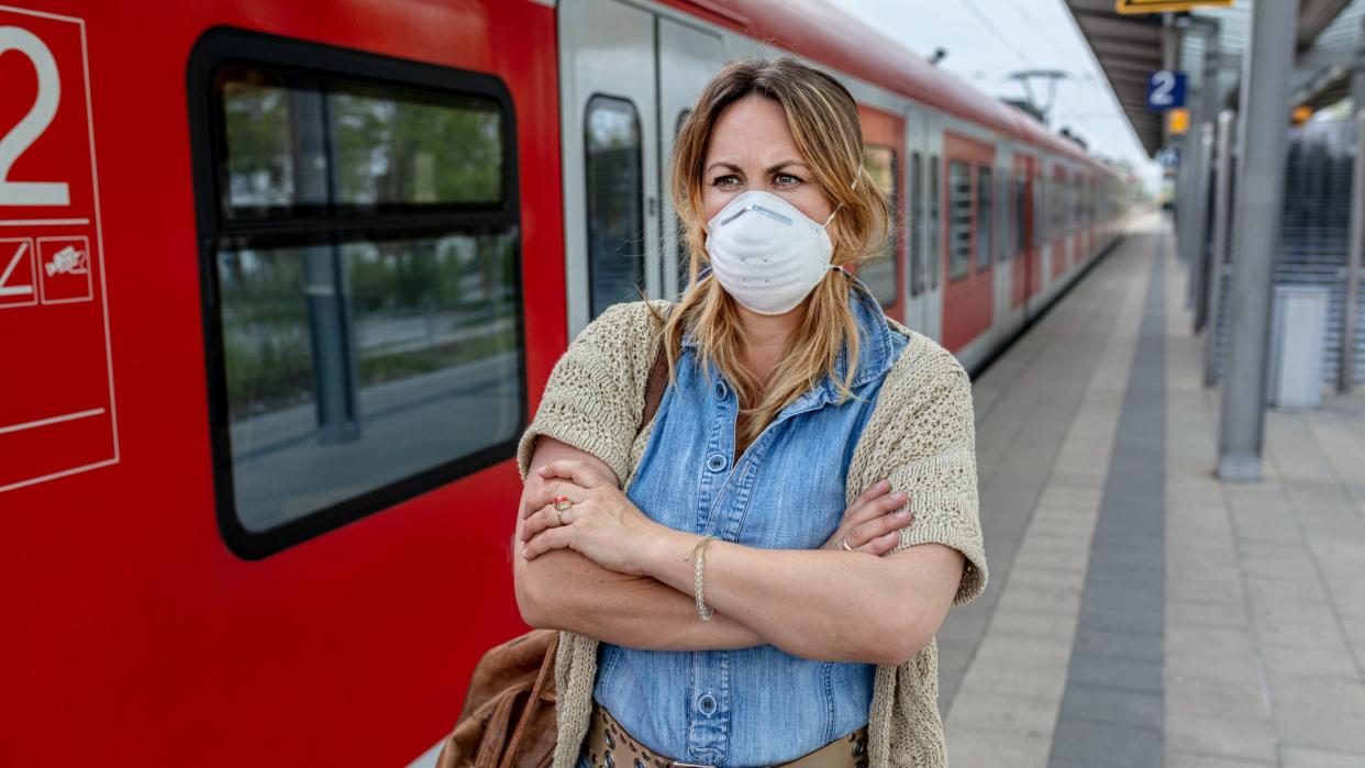  Woman wearing a protective face mask while standing on train platform with her arms crossed, looking annoyed. 