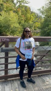 A woman in a white T-shirt and black pants on a bridge over a creek, smiling.