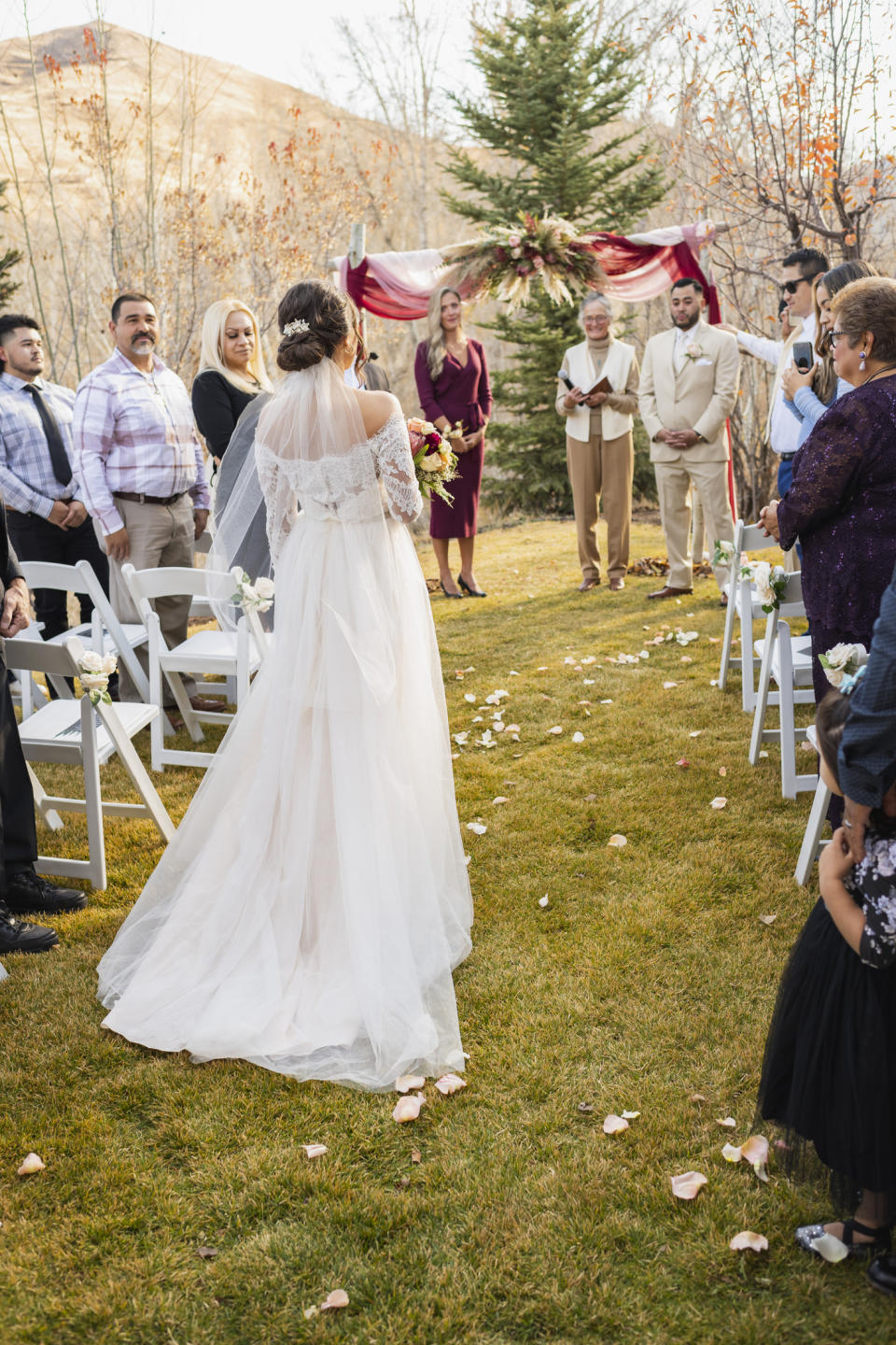 A bride in a flowing white dress holding a bouquet of flowers walks down the aisle with guests on either side during an outdoor wedding ceremony