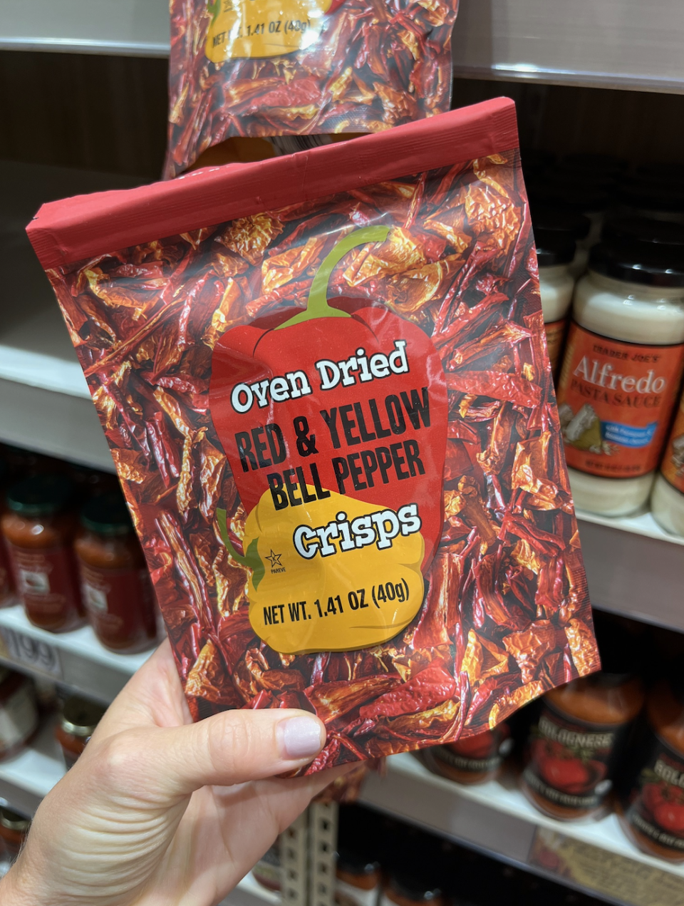 Hand holding a bag of oven-dried red and yellow bell pepper crisps (net weight 1.41 oz) in a grocery store aisle with various pasta sauces on shelves