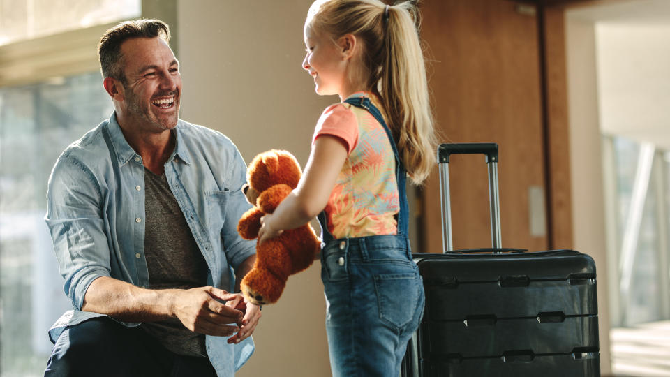 A man smiles while speaking to a young girl holding a teddy bear. A suitcase with wheels stands next to them indoors