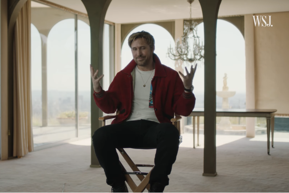 Man in red jacket and white shirt sits on a chair, gesturing with hands, indoors with arched windows