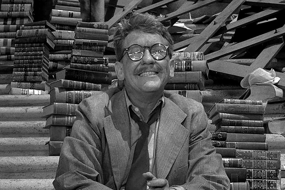 A man in glasses smiles in front of stacks of books.