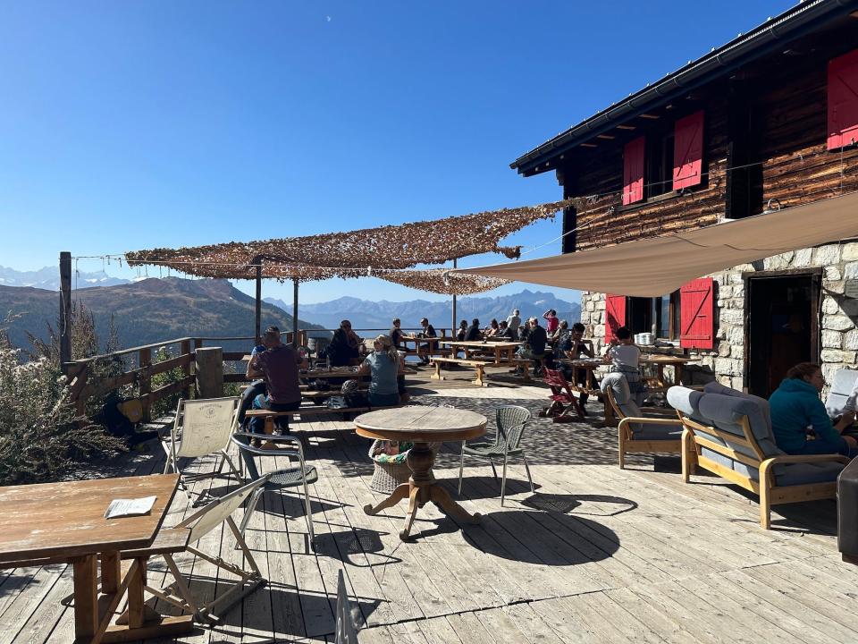 wooden porch with people sitting in assorted tables chairs drinking and eating next to wood stone house with mountains in background