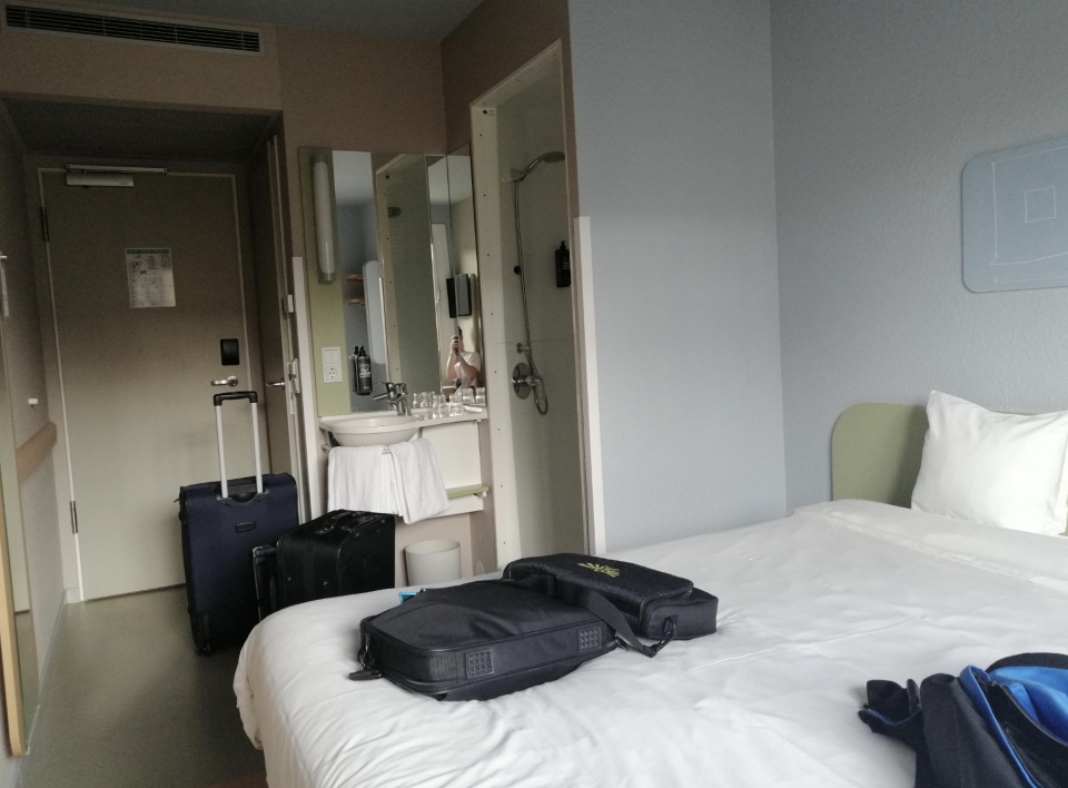 Hotel room with a bed, suitcase, and bags. A bathroom with a sink and mirror is visible