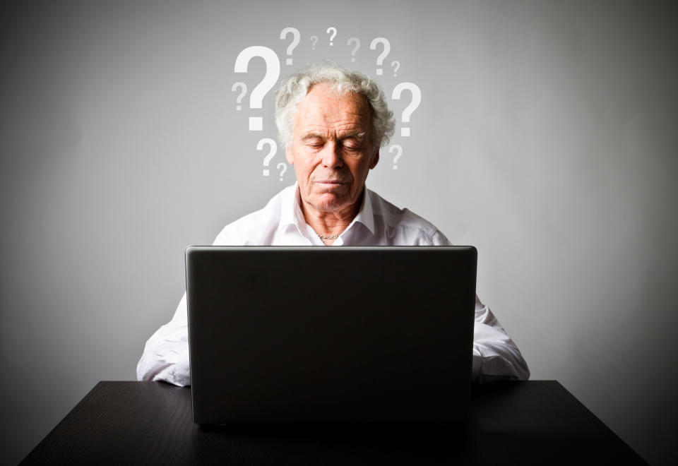 A senior man working on a laptop in front of a wall with question marks drawn on it.