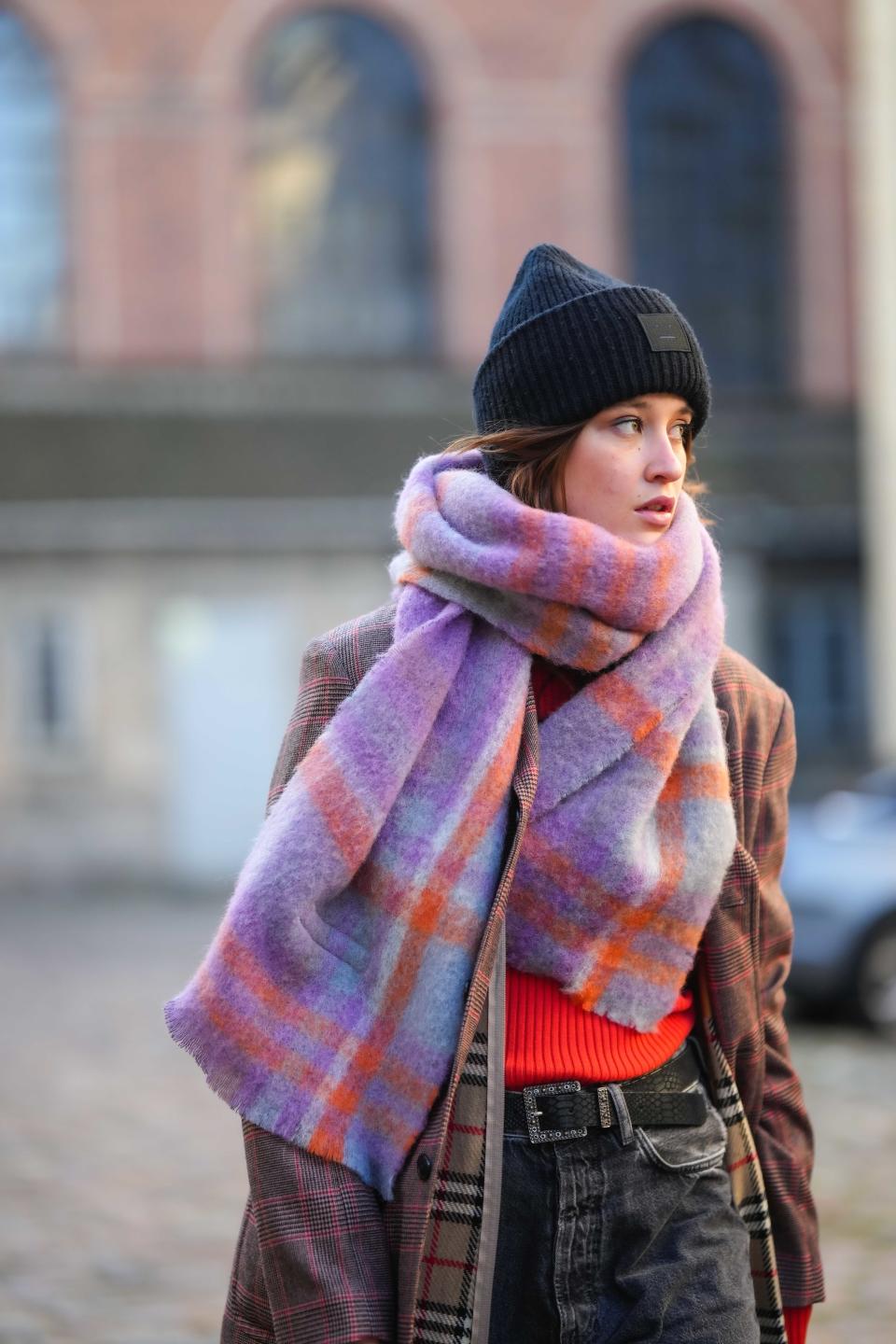 A person is standing outside, wearing a beanie, a plaid coat, a colorful scarf, a red sweater, and jeans. They look to the side with a contemplative expression