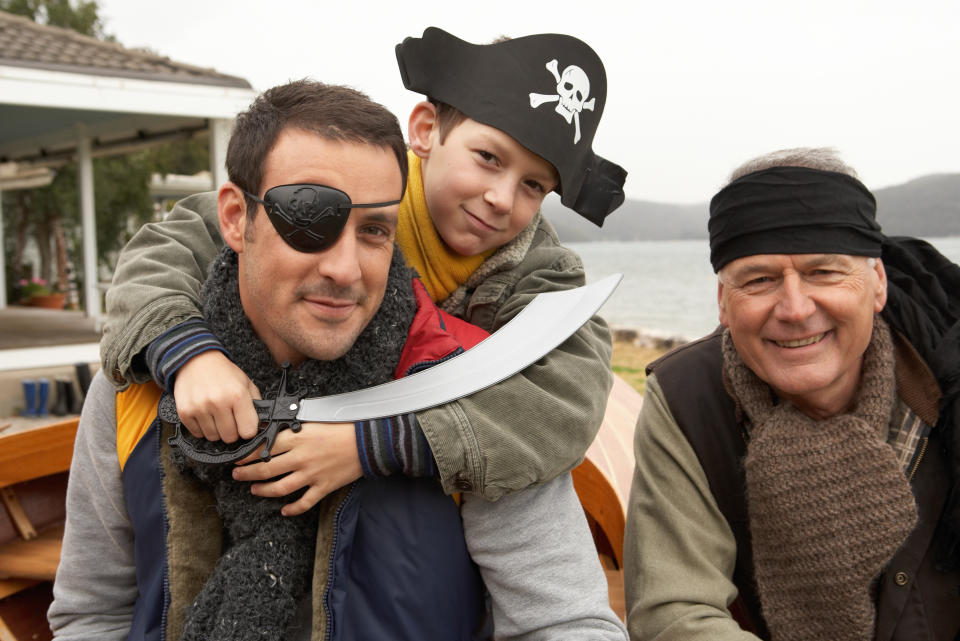 Three people dressed as pirates, including a young boy with a toy sword and pirate hat with a skull and crossbones. The setting is outdoors by a body of water