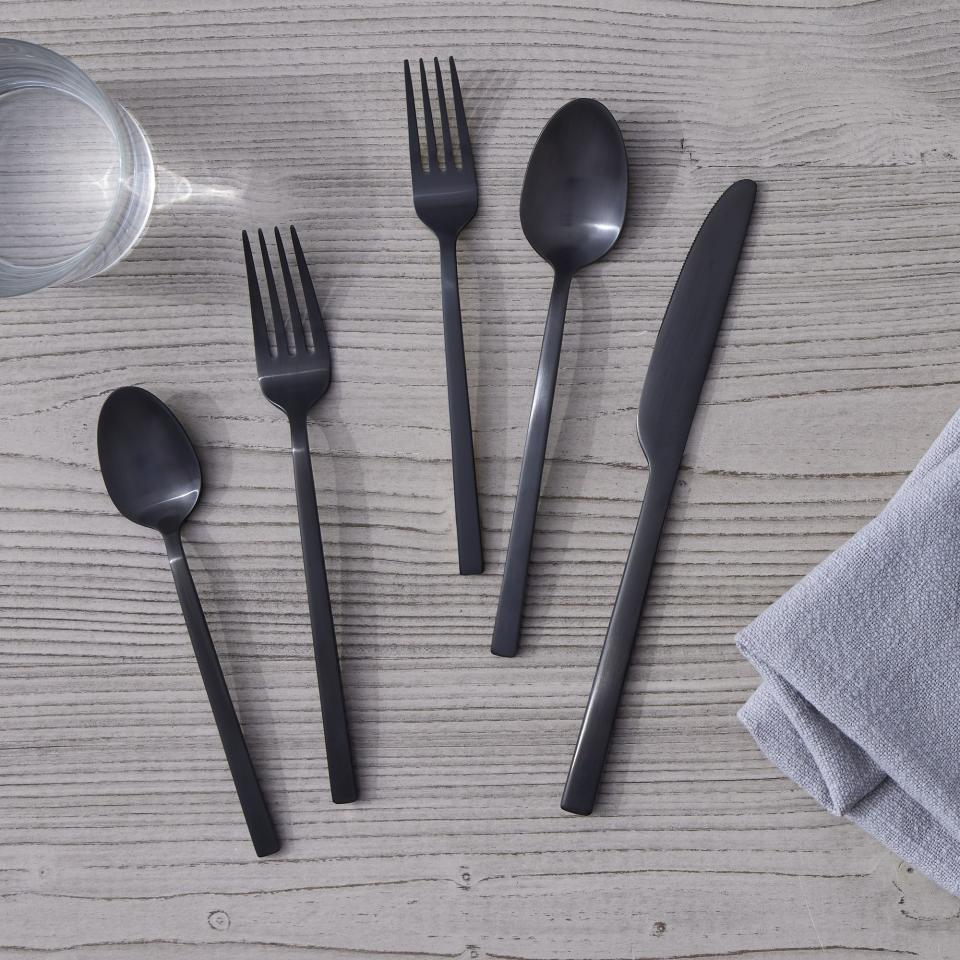 Five pieces of black silverware on light grey wood table with grey napkin and water glass