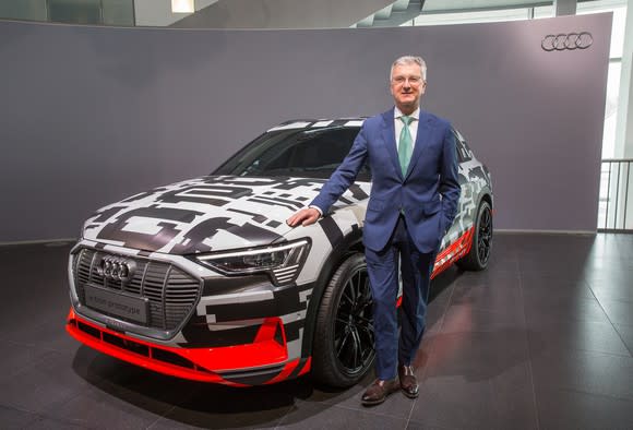 Stadler, in a blue suit, is standing next to the Audi etron quattro prototype, a midsize electric SUV wrapped in black and white camouflage.