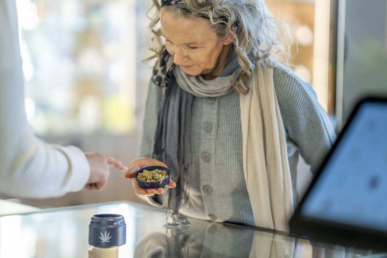 A mature woman stands at the counter of a legal cannabis retailer as she holds out a lid of buds.  She is dressed casually and looking down to inspect the product as the associate tries to sell it to her.