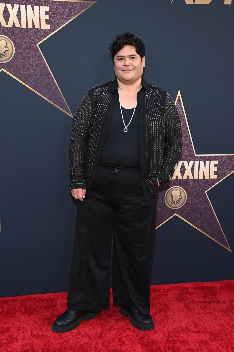 Harvey Guillén on the red carpet in a stylish outfit with a sheer, striped jacket and a silver necklace