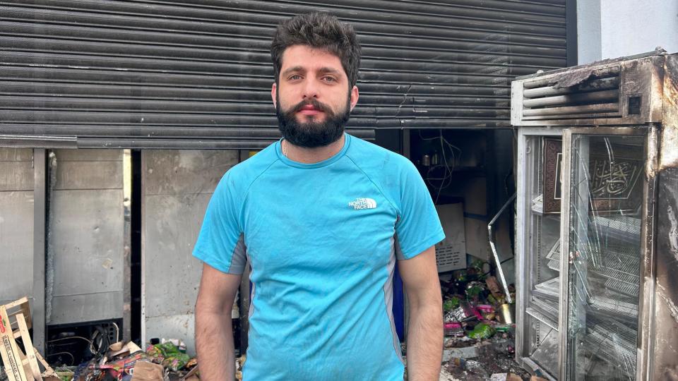 Jamal Ghabes, wearing a blue T-shirt, stands in front of a burned-out shop