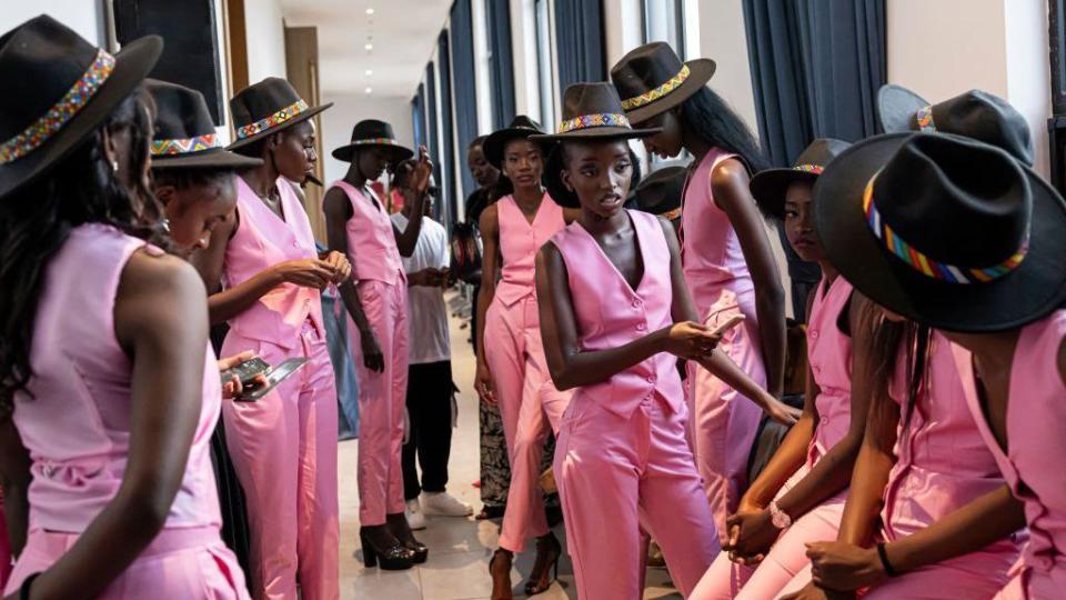 Contestants in matching pink suits and black hats wait backstage.