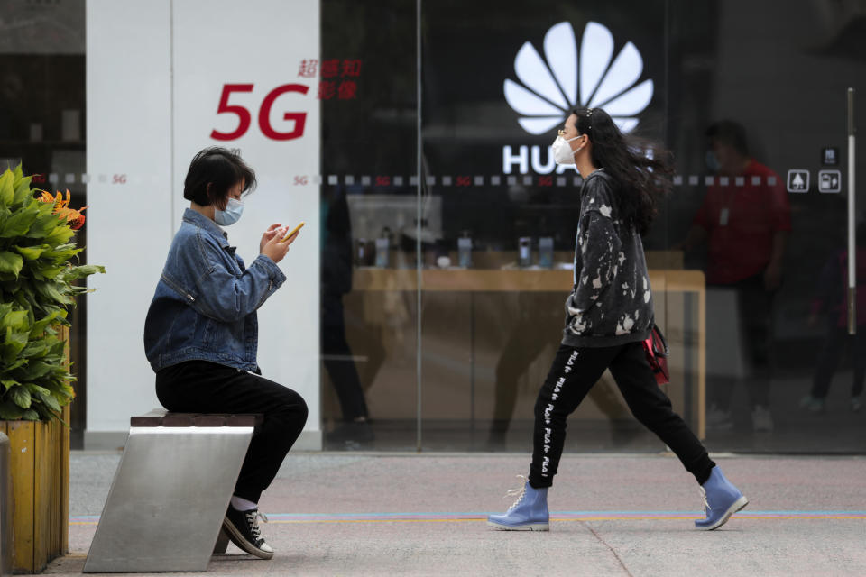 FILE - In this Oct. 11, 2020, file photo, a woman wearing a face mask to help curb the spread of the coronavirus browses her smartphone as a masked woman walks by the Huawei retail shop promoting it 5G network in Beijing. Chinese tech giant Huawei said Wednesday, March 31, 2021, it eked out a gain in sales and profit last year but growth plunged after its smartphone unit was hammered by U.S. sanctions imposed in a fight with Beijing over technology and security. (AP Photo/Andy Wong, File)