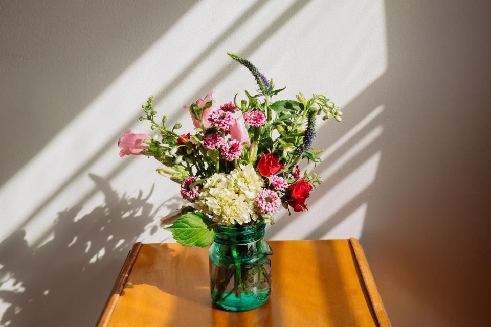 A vibrant bouquet of mixed flowers in a green vase sits on a wooden table, with sunlight casting diagonal shadows on the wall behind