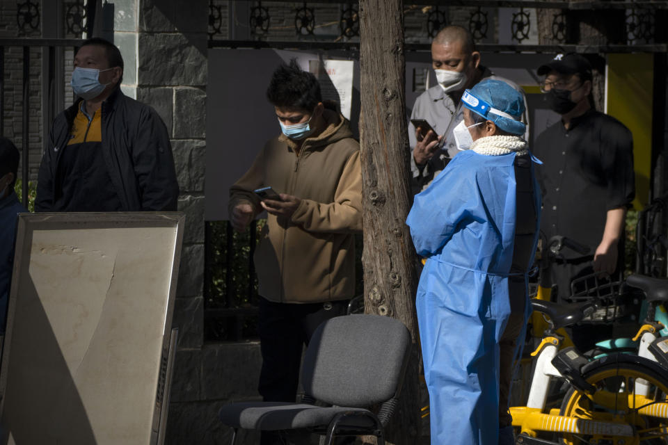 A worker wearing a protective suit watches as people line up for COVID-19 tests at a coronavirus testing site in Beijing, Saturday, Nov. 5, 2022. (AP Photo/Mark Schiefelbein)