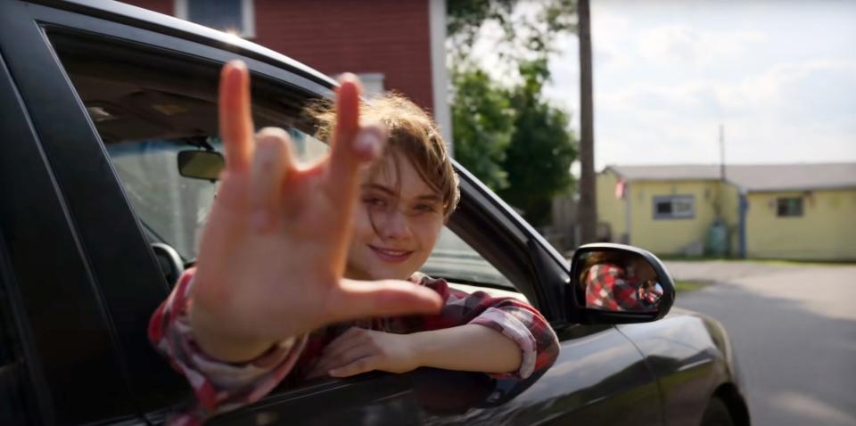 A white girl with brown hair sits in the passengers seat of a car. She looks back out the window signing, "I love you" in sign laungage.