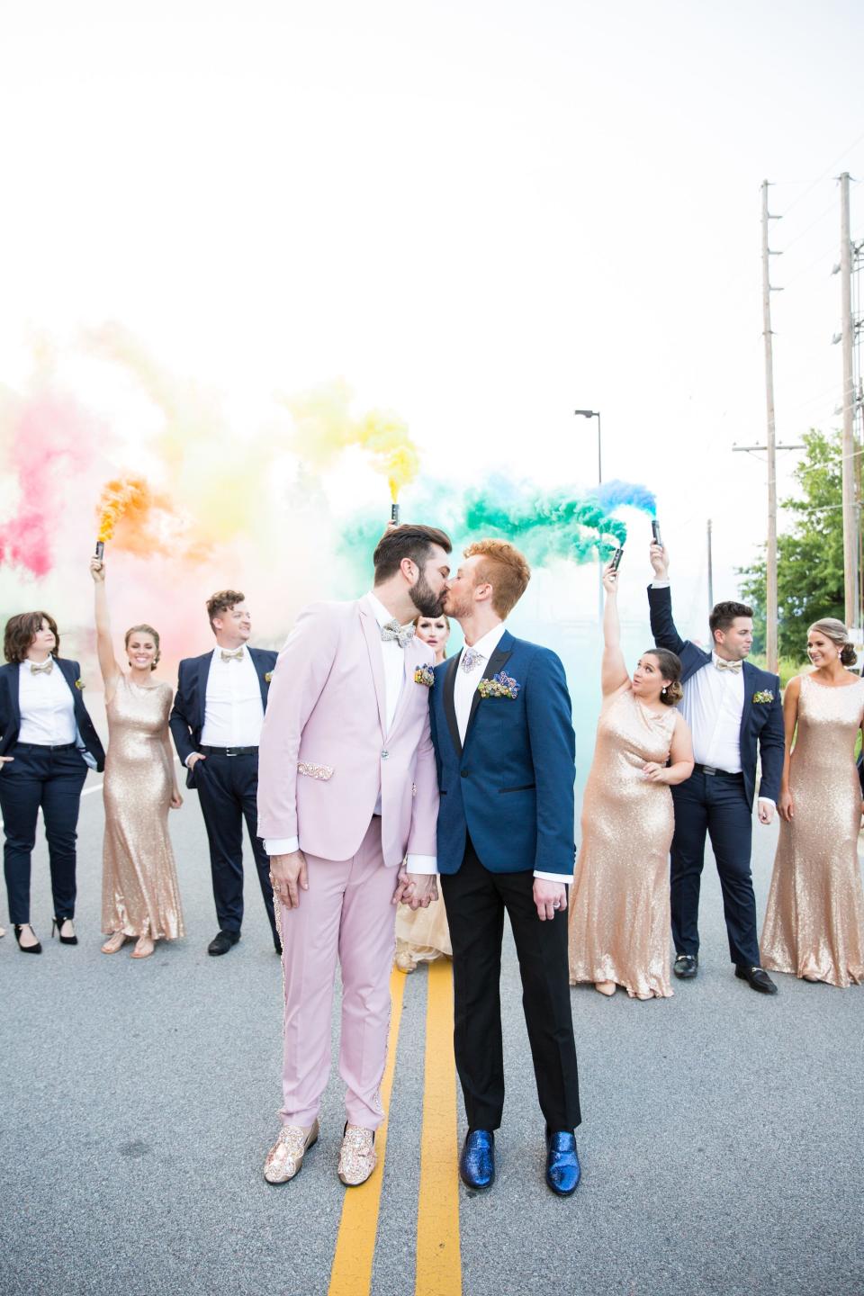 Two grooms kiss as their wedding party holds colorful smoke bombs behind them.