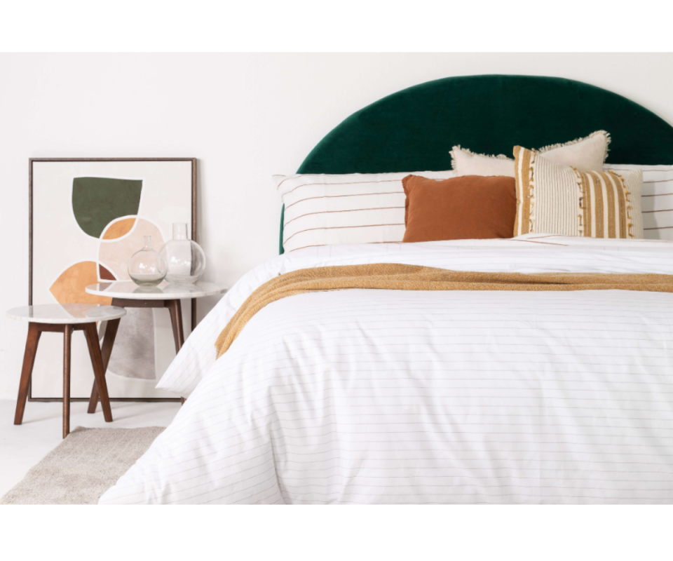 A white bed sits on a pale floor with orange patterned cushions infront of a deep green velvet arched bedhead. 