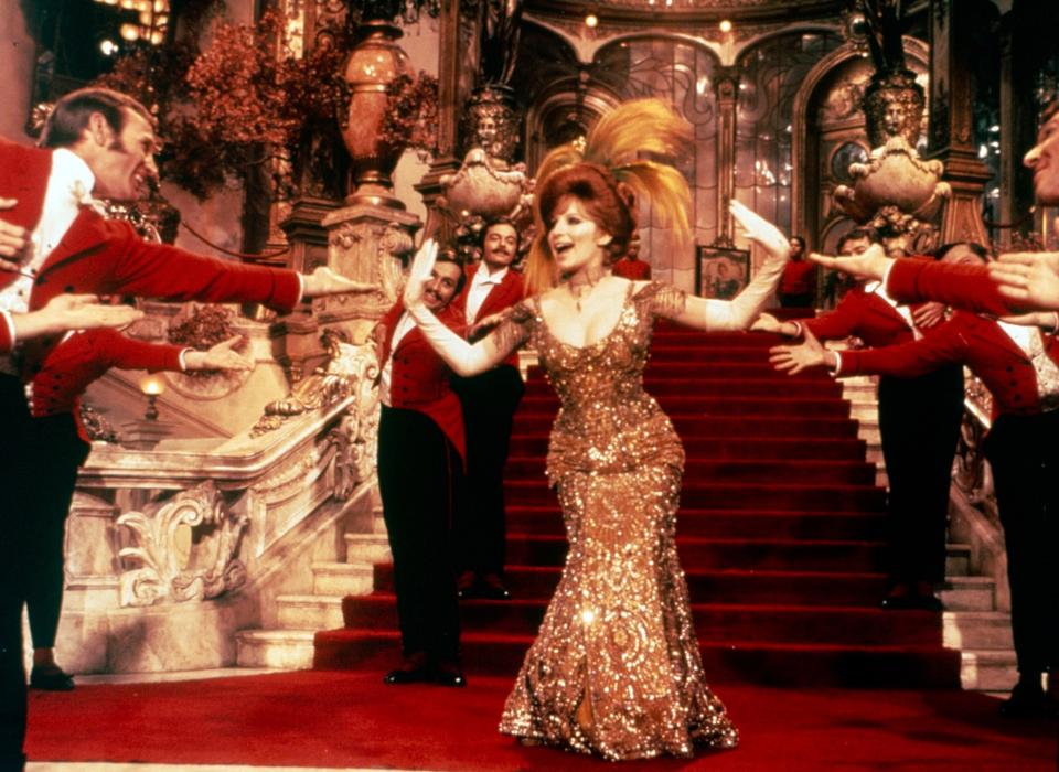 Barbra Streisand as Dolly sings in front of a large, red-carpeted staircase