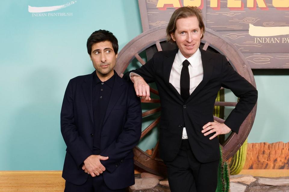 jason schwartzman, wearing a black suit, looks to the side and crosses his hands, as wes anderson wears a black suit and tie and rests his arm on schwartzman's shoulder