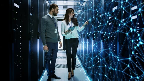 A man and woman standing in an aisle of large computer servers with a web of bright blue dots overlaying the servers the woman is point to.