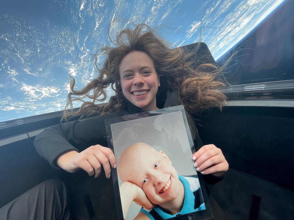 a woman with red hair poses next to a window through which Earth can be seen. she is holding a picture of herself as a young child with no hair