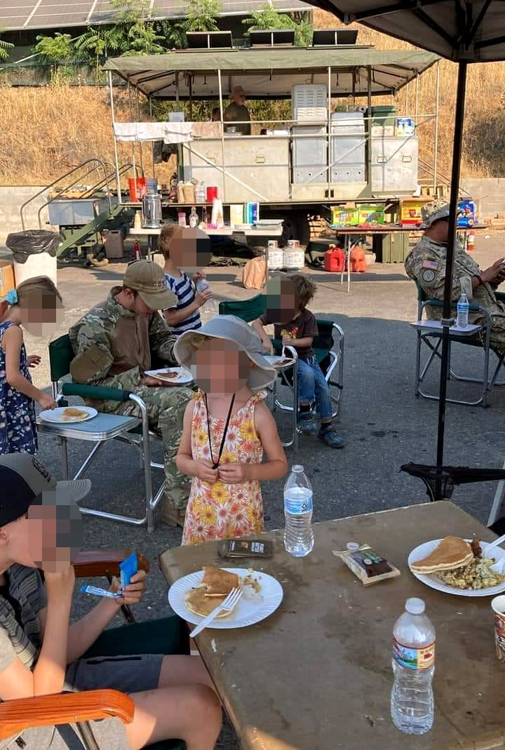 Image: Militia members in military fatigues eat breakfast in the parking lot of H&L Lumber in Mariposa, Calif., on July 24, 2022. Children's faces have been obscured by NBC News. (via Facebook)
