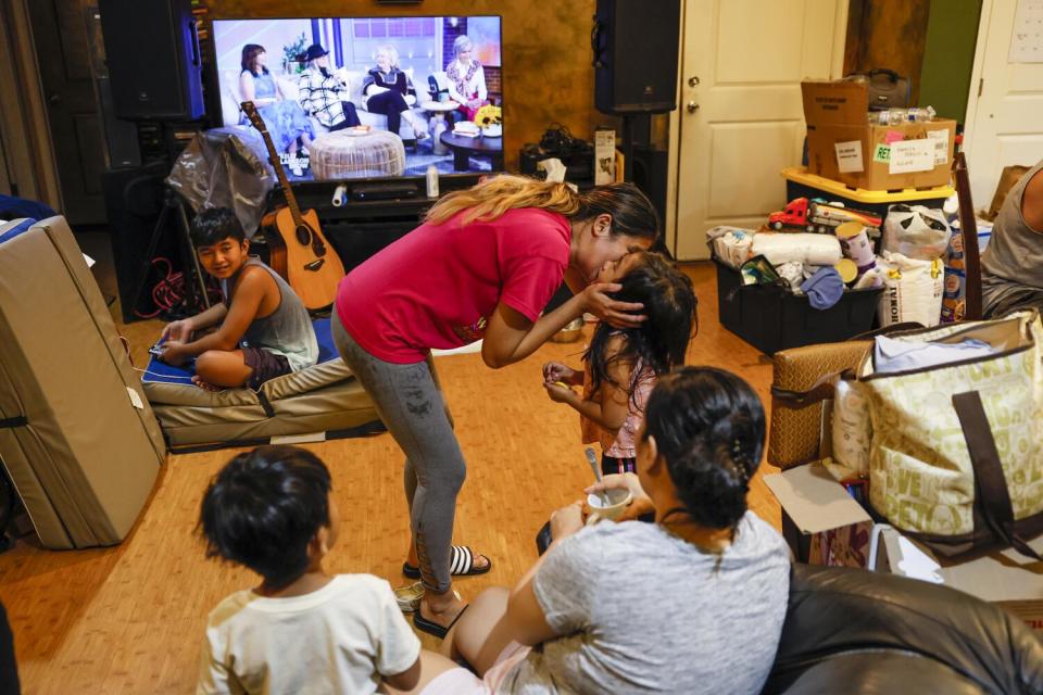 A woman in a red shirt kisses a young girl on the cheek, surrounded by other people, with the TV on the background