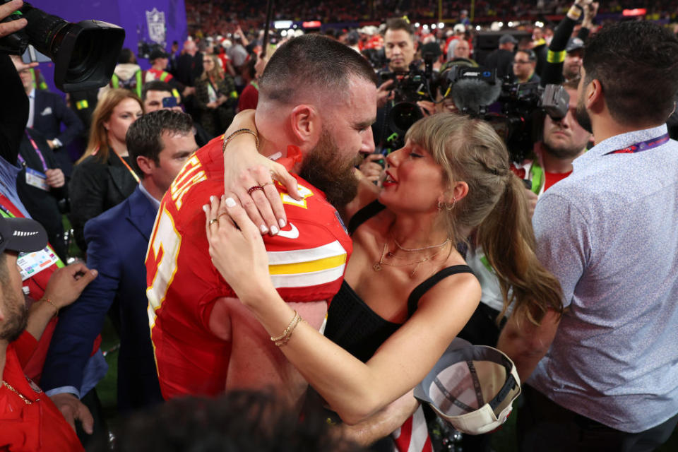 Travis Kelce in football gear and Taylor Swift in a sleeveless black outfit share a celebratory hug among a crowd at a sports event