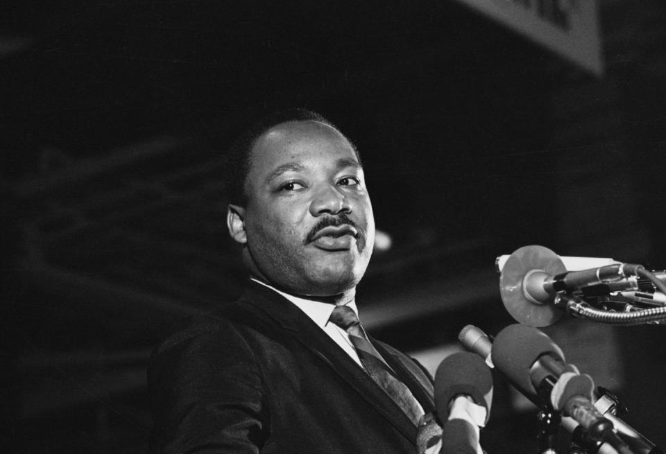 martin luther king jr speaks into several microphones and looks to the left, he wears a suit and tie